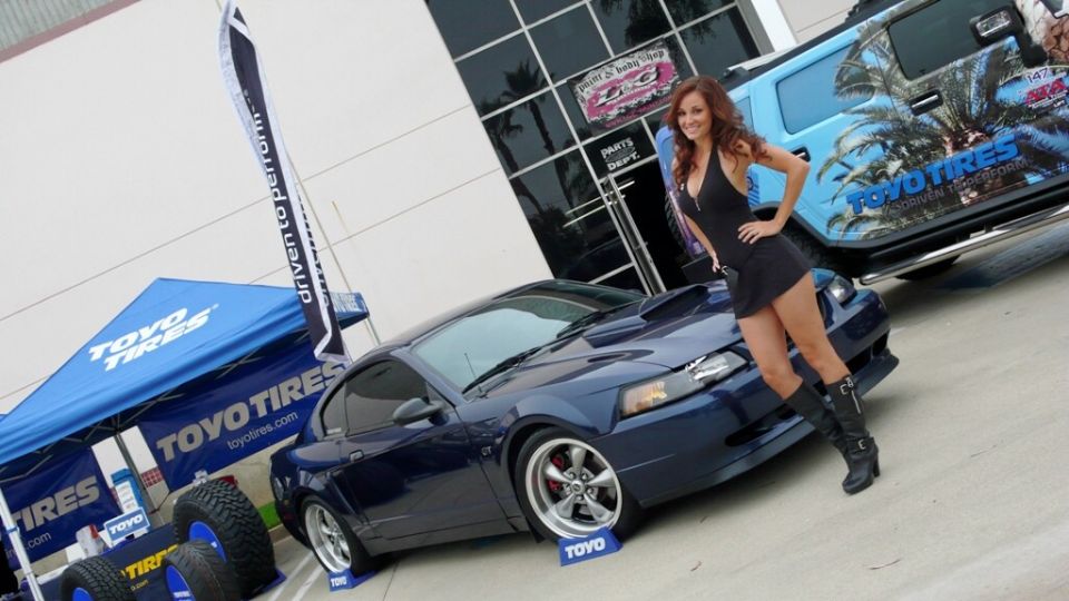 Dark blue 2001 Ford Mustang coupe with hood scoop, parked on display with a woman posing. A blue tent and a light blue SUV are also visible