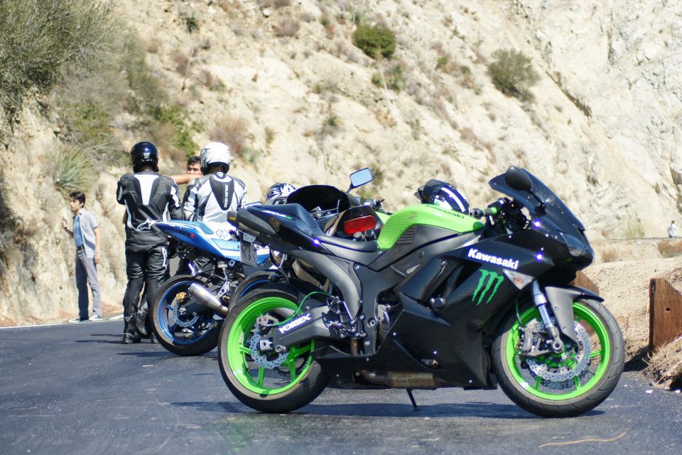 Two people in motorcycle gear stand next to a black and green Kawasaki motorcycle. Another blue motorcycle is behind them