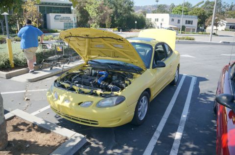 1995 Ford Taurus - A yellow coupe with its hood and trunk open is parked in a lot next to another vehicle.
