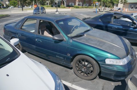 1995 Ford Taurus - A green 1995 Ford Taurus sedan with a matte gray hood is parked between two other vehicles.