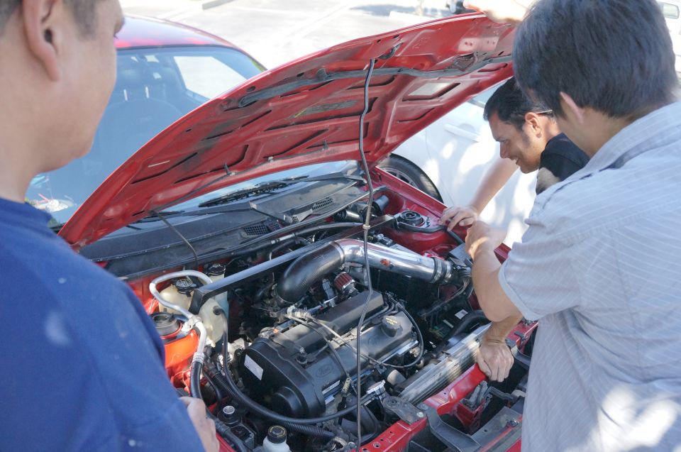 Red car engine bay with aftermarket intake, viewed from above with people gathered around
