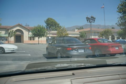 1995 Ford Taurus - Two cars are parked in a lot, with a white car to the left and buildings behind them.