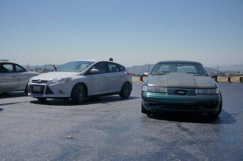 1995 Ford Taurus - A green 1995 Ford Taurus with a grey hood is parked on a paved surface under a clear sky.