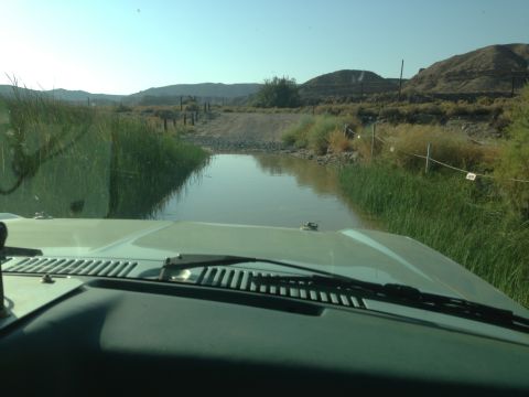 1986 Ford Bronco - The vehicle approaches a water obstacle on a dirt road, with dry, arid hills in the background.