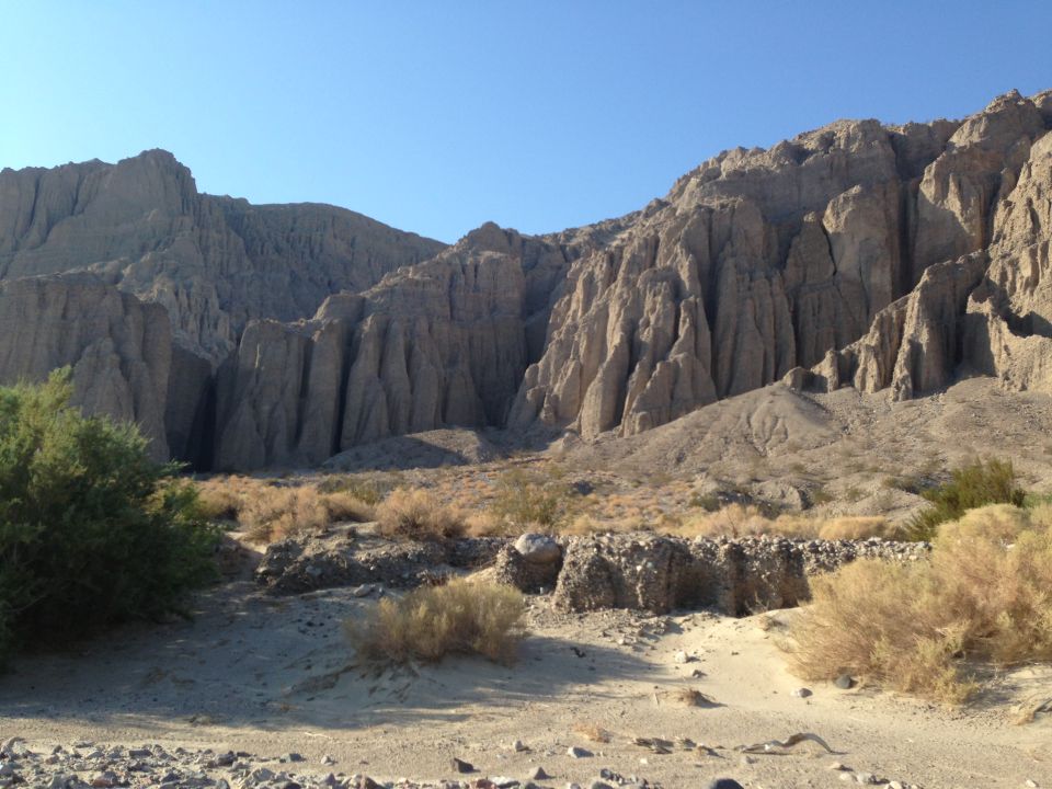 Desert canyon with eroded rock walls and sparse vegetation under a clear blue sky. A watermark reads SUPERMOTORS