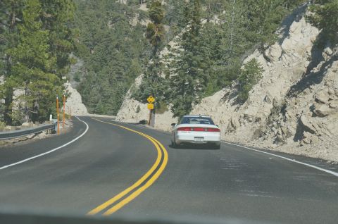 1995 Ford Taurus - A white sedan navigates a scenic mountain highway, with steep, rocky terrain and dense pine forests flanking the road.