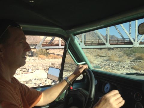 1986 Ford Bronco - The driver navigates a rough terrain, passing under a large bridge structure in a desert landscape.