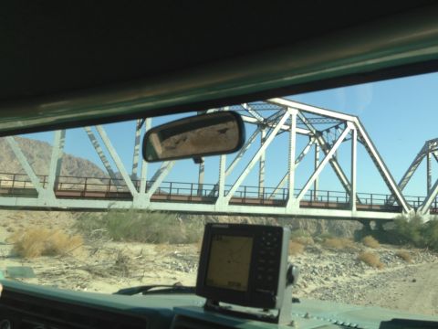 1986 Ford Bronco - View from inside a vehicle, showing a GPS unit on the dashboard as it approaches a large truss bridge in a desert environment.