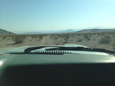 1986 Ford Bronco - View from inside a vehicle driving on a dirt road through a desert with mountains in the distance.