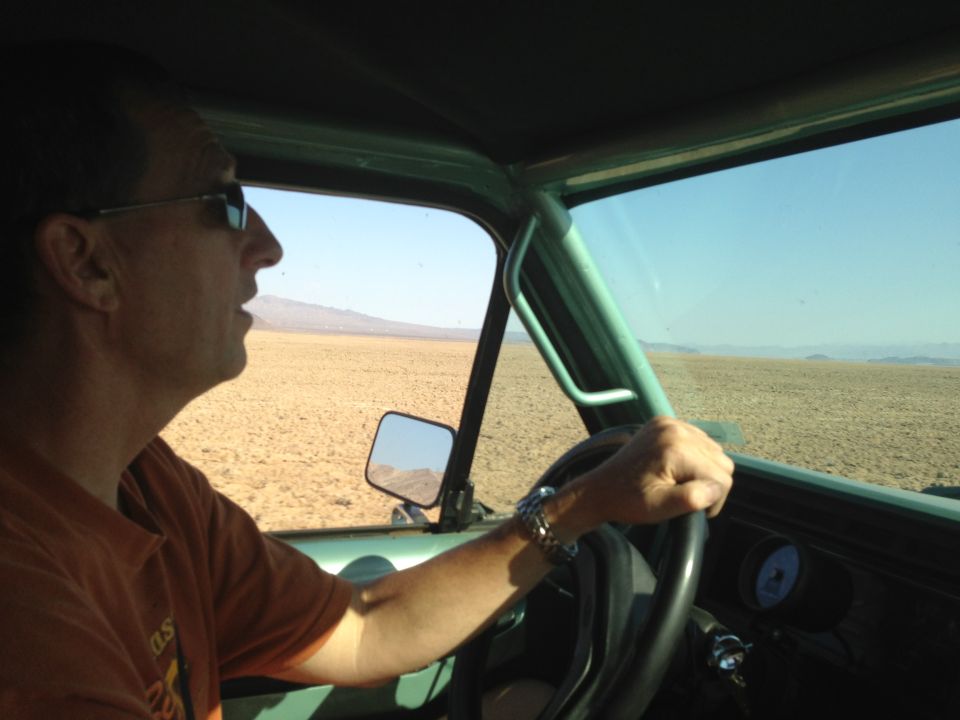 Man in sunglasses driving a green off-road vehicle through a desert landscape