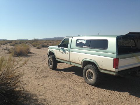 1986 Ford Bronco - A lifted, two-tone pickup truck with a camper shell sits on a dusty desert road under a clear blue sky.