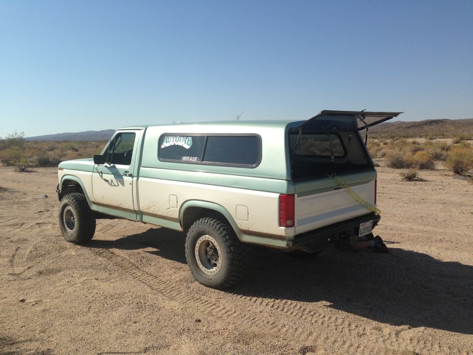 White and teal Ford F-Series pickup truck with a camper shell, lifted suspension, and off-road tires parked on a dirt road