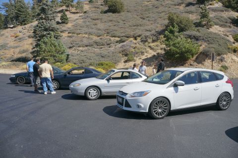 1995 Ford Taurus - A white hatchback is parked in a lot with other vehicles and people nearby.