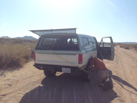 1986 Ford Bronco - A person is attending to the rear tire of a two-tone Ford Bronco, which has a camper shell and an extended awning, in a dry, open area.