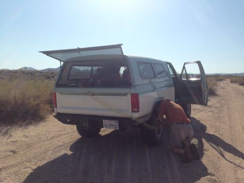 1986 Ford Bronco - A person works on the rear tire of a two-tone Ford Bronco with a camper shell on a dirt road.