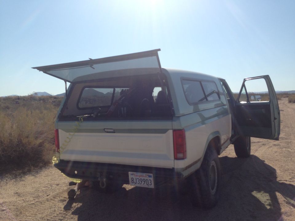 White and green Ford Bronco with camper shell, rear view, open rear hatch and driver door, desert setting
