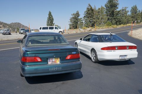 1995 Ford Taurus - Two Ford vehicles are parked on the side of a road with trees and mountains in the background.