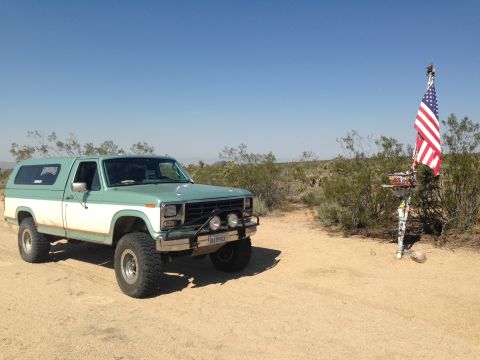 1986 Ford Bronco - A lifted, two-tone pickup truck with a camper shell sits in a desert landscape next to an American flag.