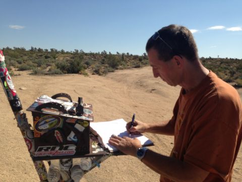 1986 Ford Bronco - A man is writing on a piece of paper in a desert environment, with a sticker-covered box to his left.