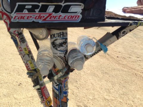 1986 Ford Bronco - A close-up of a metal structure adorned with stickers and holding several water bottles, set against a sandy background.