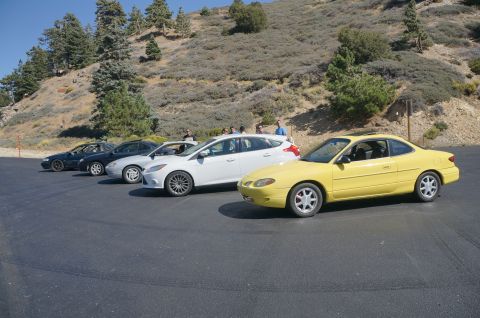 1995 Ford Taurus - A group of cars are parked in a line on a sunny day with a hillside covered in trees and brush behind them.