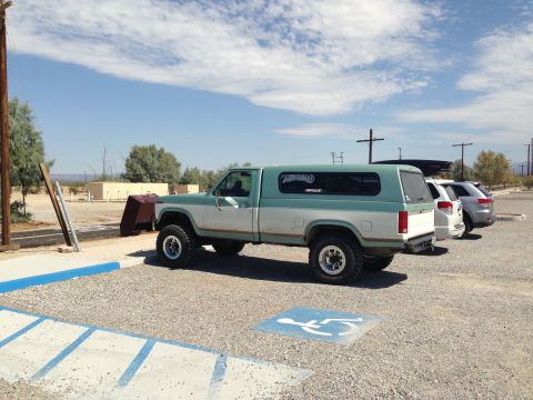 1986 Ford Bronco - A lifted two-tone green and white pickup truck with a camper shell is parked in a lot next to other vehicles.