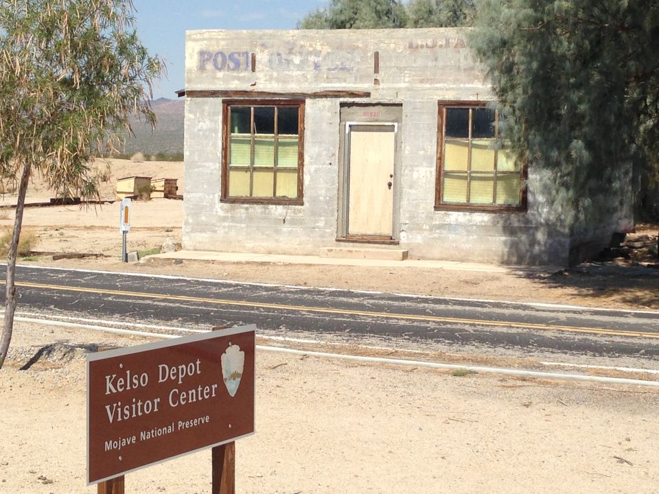 Abandoned concrete building with 'POST' painted on top, next to a road and a sign for Kelso Depot Visitor Center