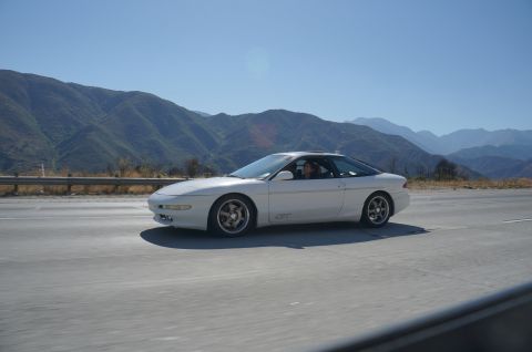 1995 Ford Taurus - A white 1995 Ford Taurus coupe is captured in motion on a highway, with a scenic mountain range visible behind it.