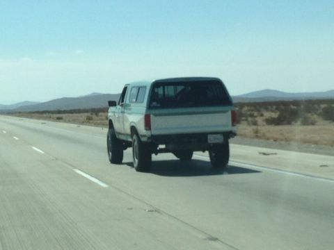 1986 Ford Bronco - A white and teal SUV with a camper shell drives down a highway in a desert landscape.