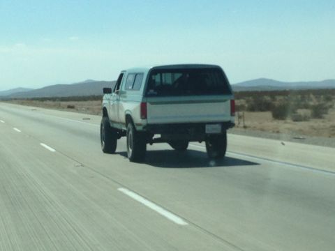 1986 Ford Bronco - A lifted SUV with a two-tone paint scheme travels down a desert highway.