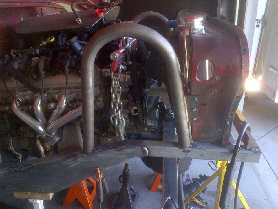 Red vehicle body with V8 engine and chrome exhaust headers, undergoing roll cage installation