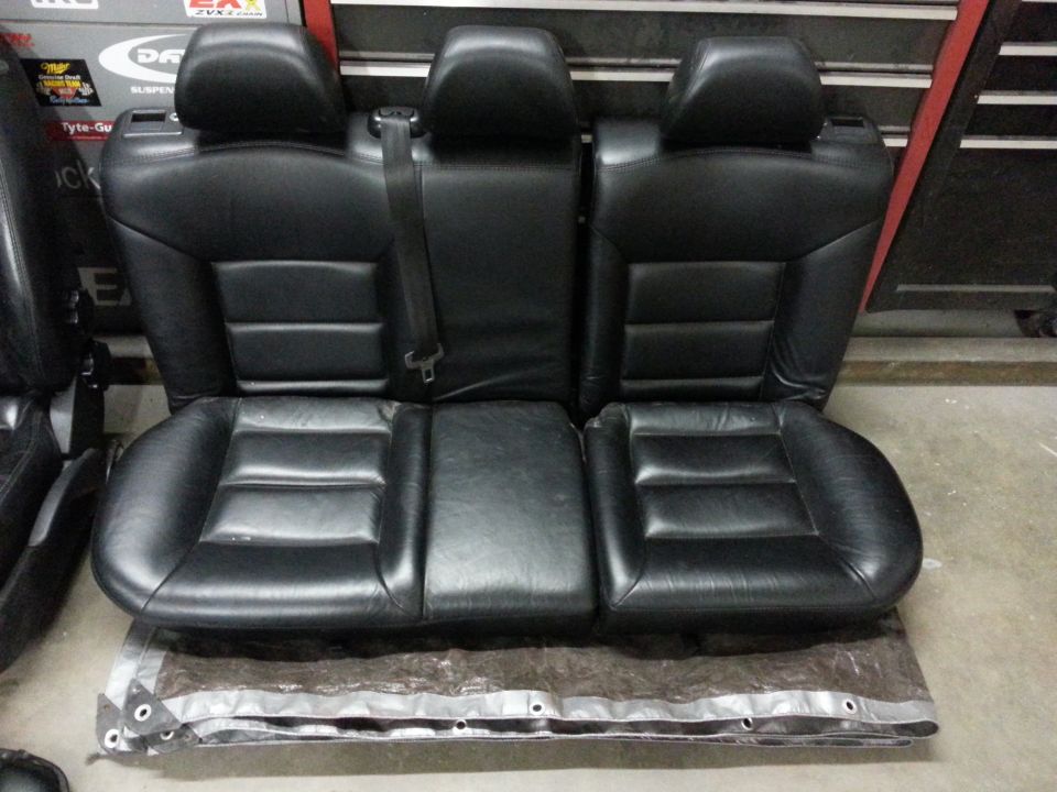 Black leather rear car seats with three headrests and seatbelts. They are on top of a tarp in a garage