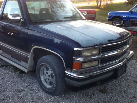 2006 Ford E350 - This dark blue pickup truck, likely from the mid-1990s, has a hood with significant paint wear. Another Chevrolet truck is parked behind it.