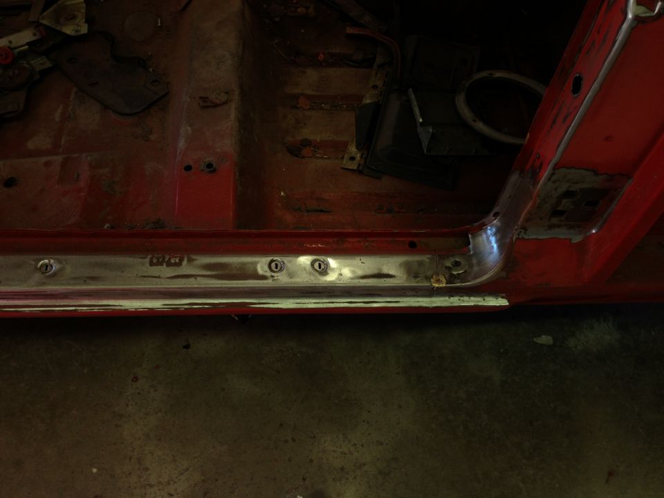Close-up of a car's red interior floor pan and door sill with visible rust and some metal trim. Text is visible on the trim