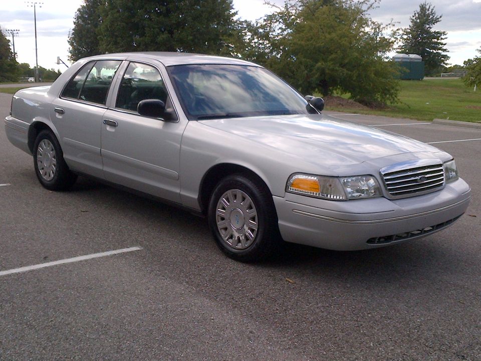 Silver 2001 Ford Crown Victoria parked on asphalt with trees and a portable toilet in the background