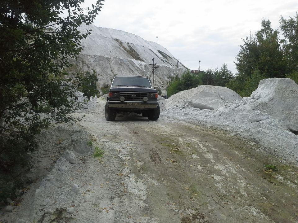 Front view of a 1987 Ford Bronco driving on a dirt road near a large pile of white material