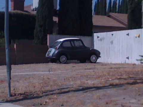 1968 Ford Mustang - A small, dark Subaru 360 is parked on a dry, grassy area next to a white fence.