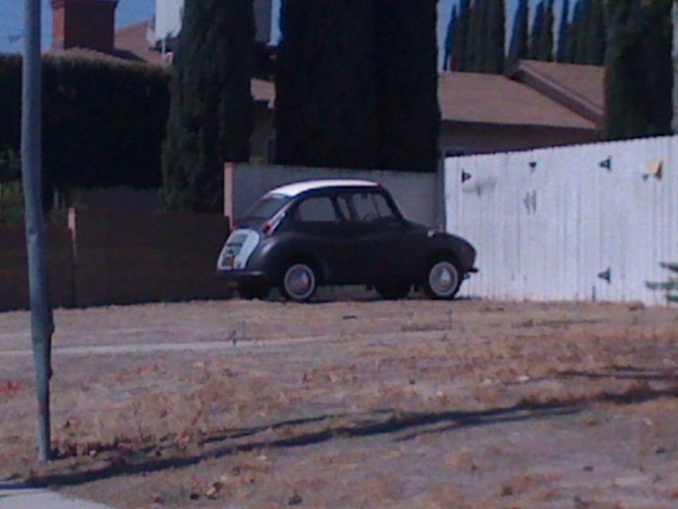 Dark Subaru 360 with white roof parked by a fence