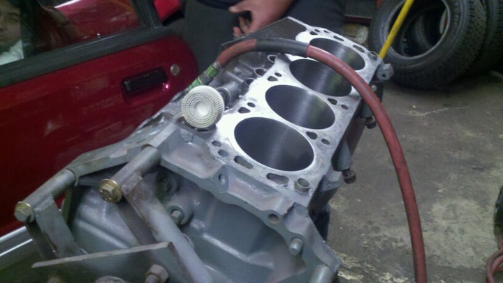 Engine block with cylinders and attached hose. Part of a red vehicle visible