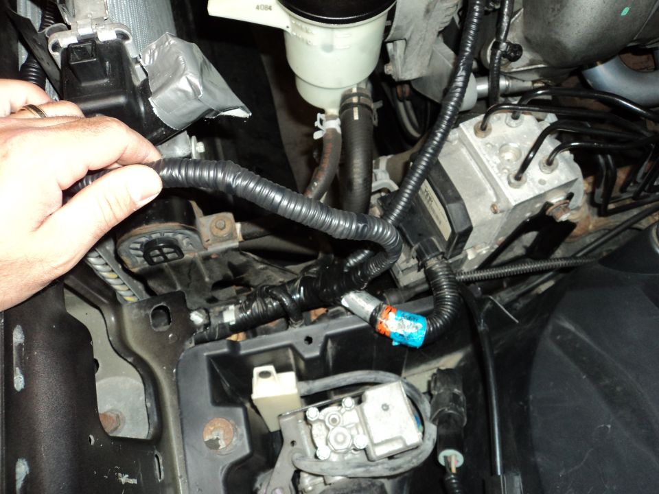 Hand working on engine bay wiring harness, showing ABS module and fluid reservoir