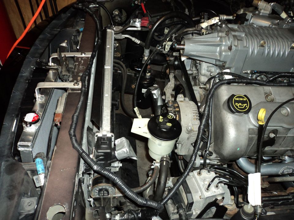 Engine bay with a silver supercharger, radiator, and various hoses. Some text is visible on engine components and a watermark