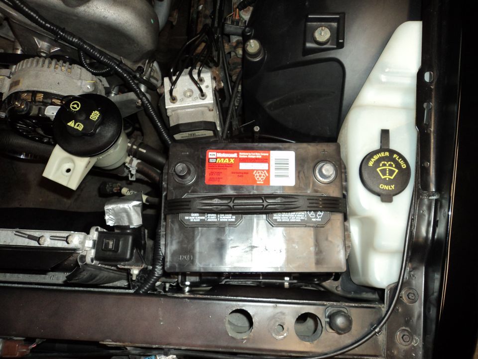 Car engine bay showing a black battery with a red label and a white washer fluid reservoir
