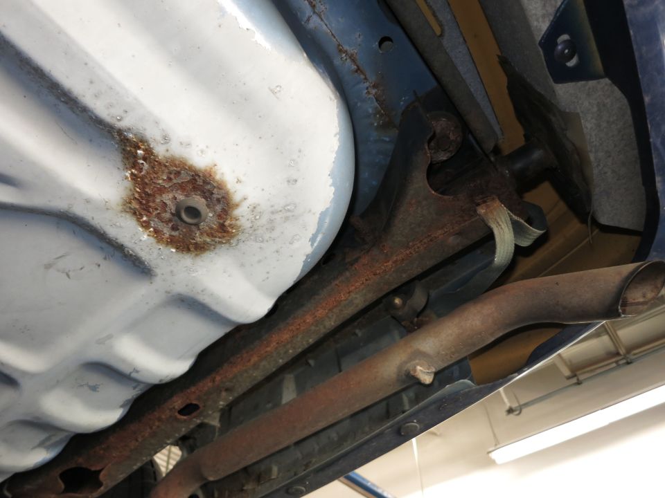 Underside view of a car's fuel tank and exhaust system, showing rust on the fuel tank and exhaust pipes