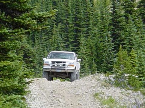 1995 Ford Bronco - A white Ford F-150 truck drives up a rocky incline on a forest road.