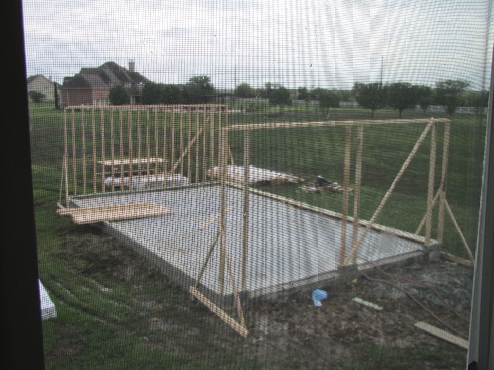 House framing is underway on a concrete slab, with lumber stacked nearby. Construction site with houses in background