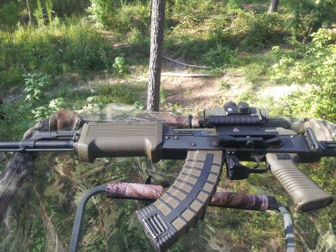 1986 Ford Bronco - AK-pattern rifle with a tan stock, mounted optic and curved magazine resting on a camouflaged surface overlooking woods.