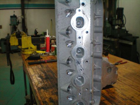 2005 Chevrolet Suburban - Close-up of an engine cylinder head resting on a wooden workbench, with various tools and industrial equipment visible in the blurred backg…