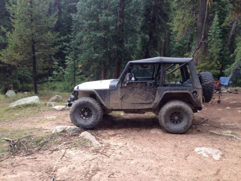 1986 Ford Bronco - A muddy Jeep Wrangler TJ sits off-road in a wooded area.