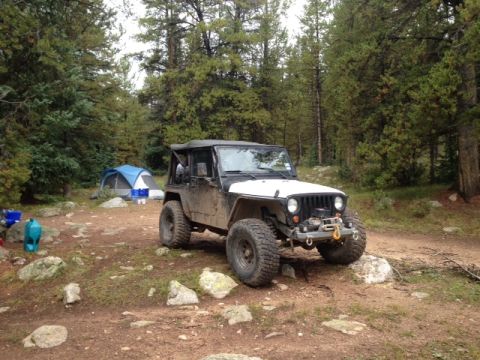 1986 Ford Bronco - A dirty Jeep Wrangler is parked on a dirt path next to a tent in a wooded area.