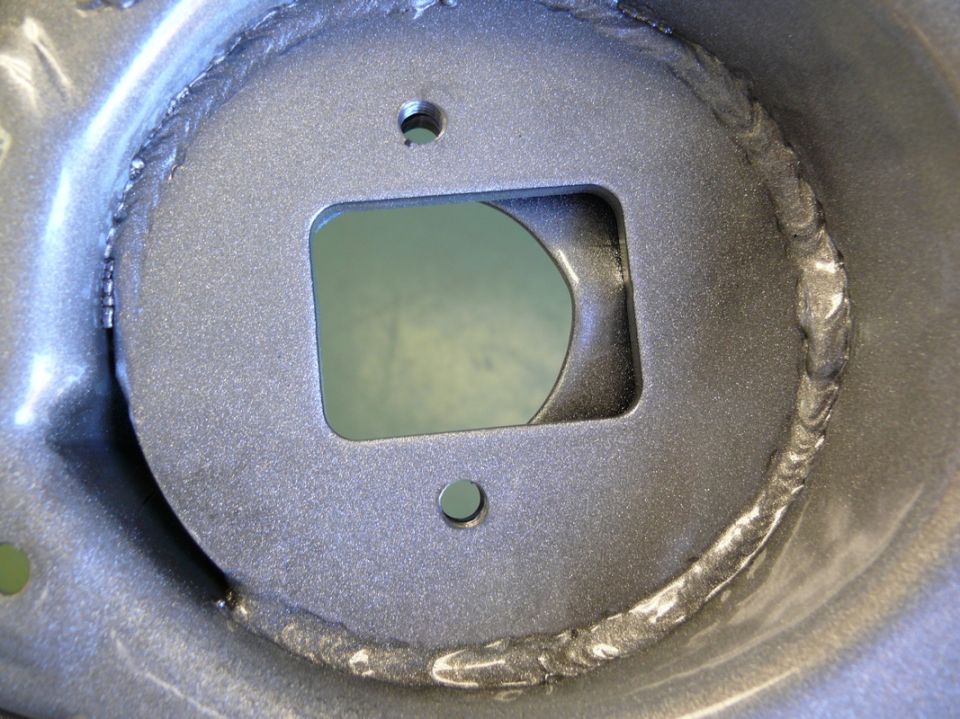Close-up of a gray metal bracket with a square cutout and two bolt holes, showing rough weld marks around the perimeter